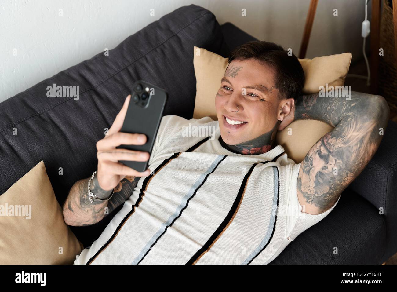 Handsome young man with tattoos smiles while enjoying his time on the phone at home. Stock Photo