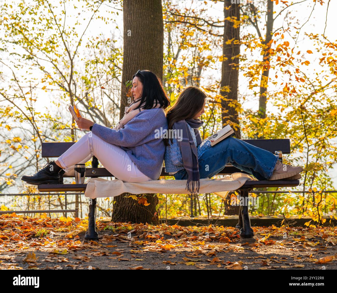 Happy mom and teenage daughter sitting on a bench in a fall park, reading a book. Family bonding, togetherness, family Stock Photo