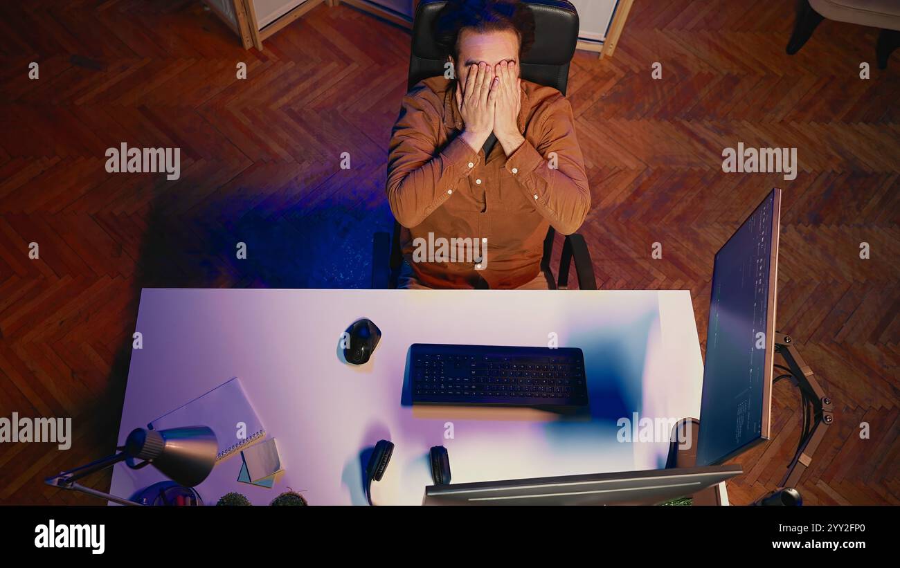 Top down view of software engineer putting face in hands in frustration while updating binary code script on computer, receiving errors. IT worker exasperated by annoying bugs while programming, aerial shot Stock Photo