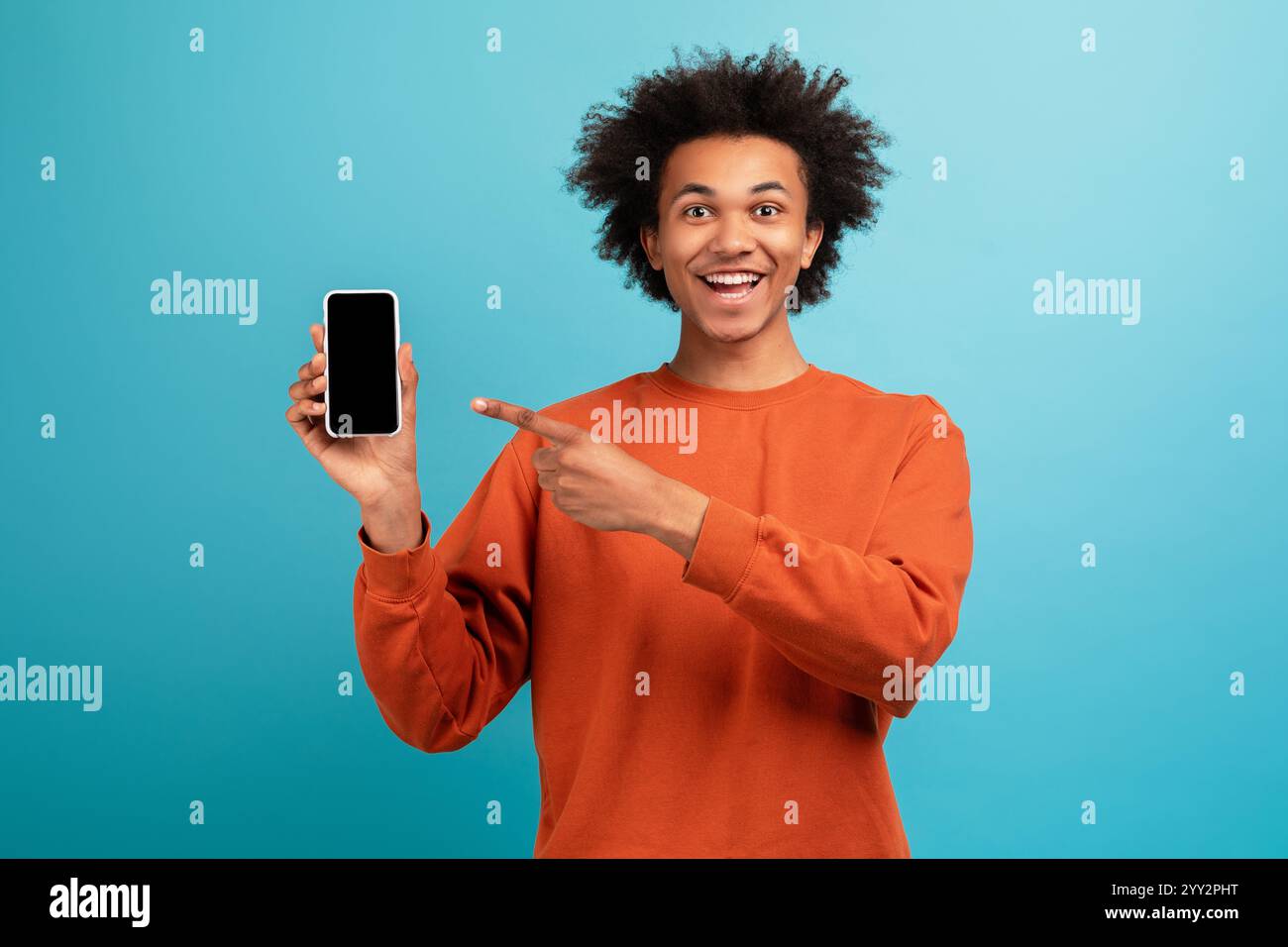 Curly-haired young man smiles and points at smartphone against blue Stock Photo
