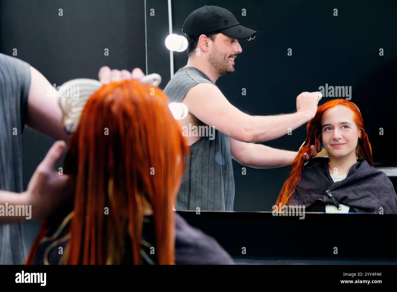A hair stylist adjusts vibrant red hair of a seated woman, reflected in a mirror at a sleek modern salon Enhance your project with this professional s Stock Photo