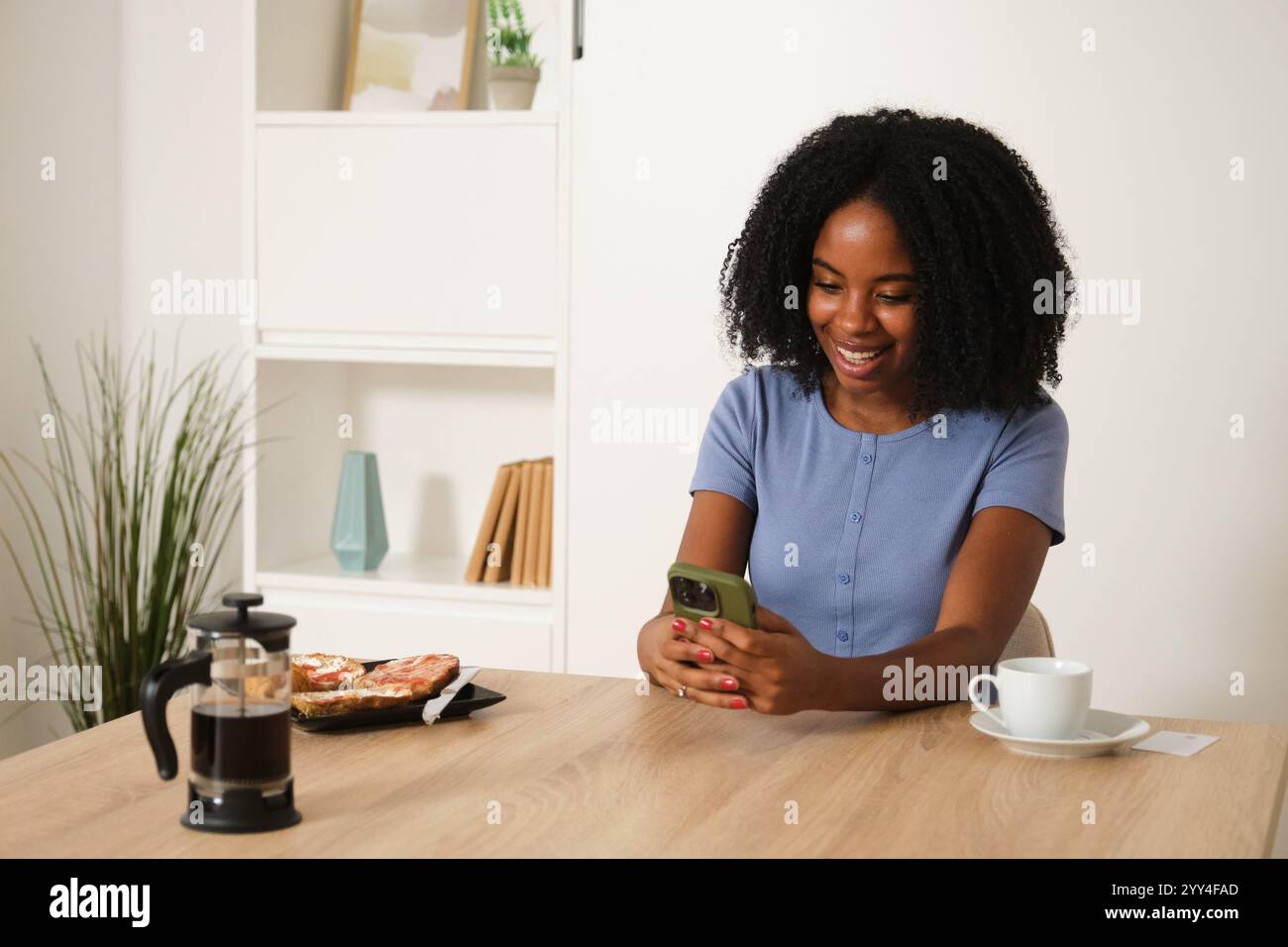 A young African American woman enjoys coffee while using her smartphone at a cozy home setting, blending relaxation with modern technology for a casua Stock Photo