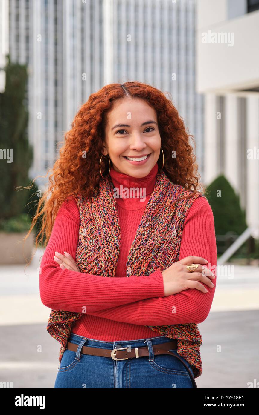 A stylish Latin woman with curly hair stands confidently in an urban setting, wearing a red sweater and colorful vest She smiles warmly, embodying a v Stock Photo