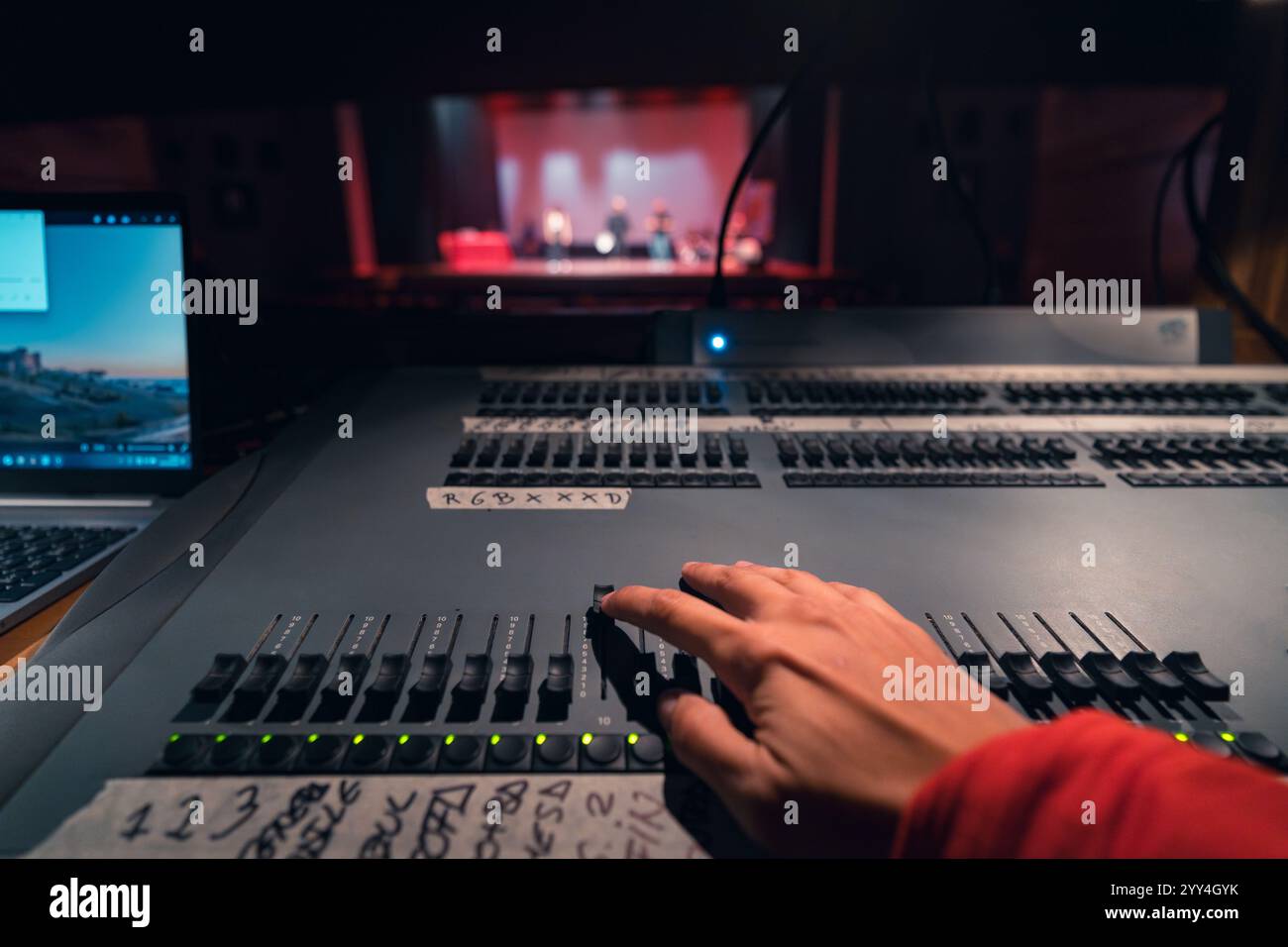 A hand adjusts a control on a theater lighting desk, enhancing stage ambiance. The stage is dimly visible in the background, showcasing a live perform Stock Photo