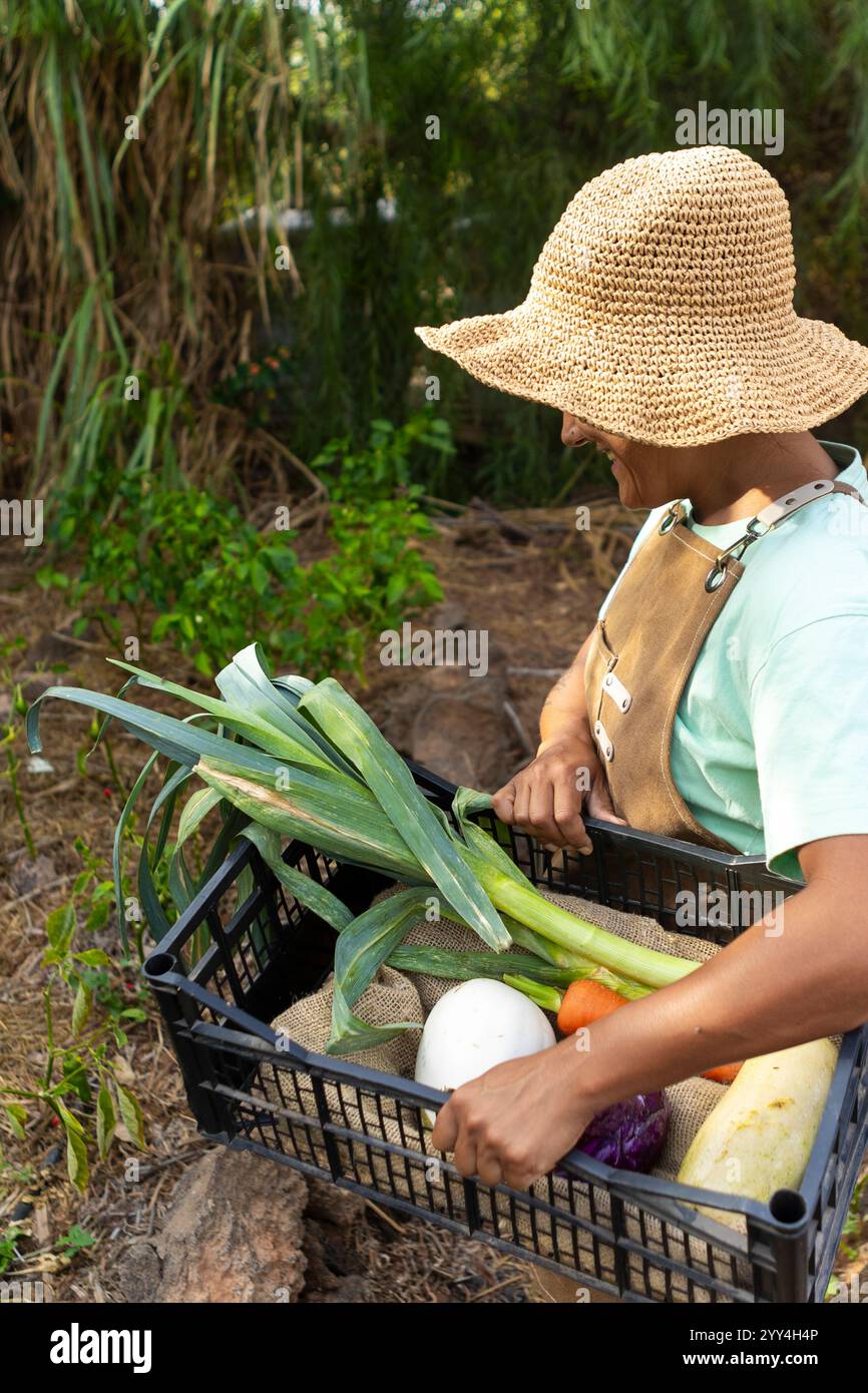 A farmer wearing a straw hat and apron carries a crate filled with ...