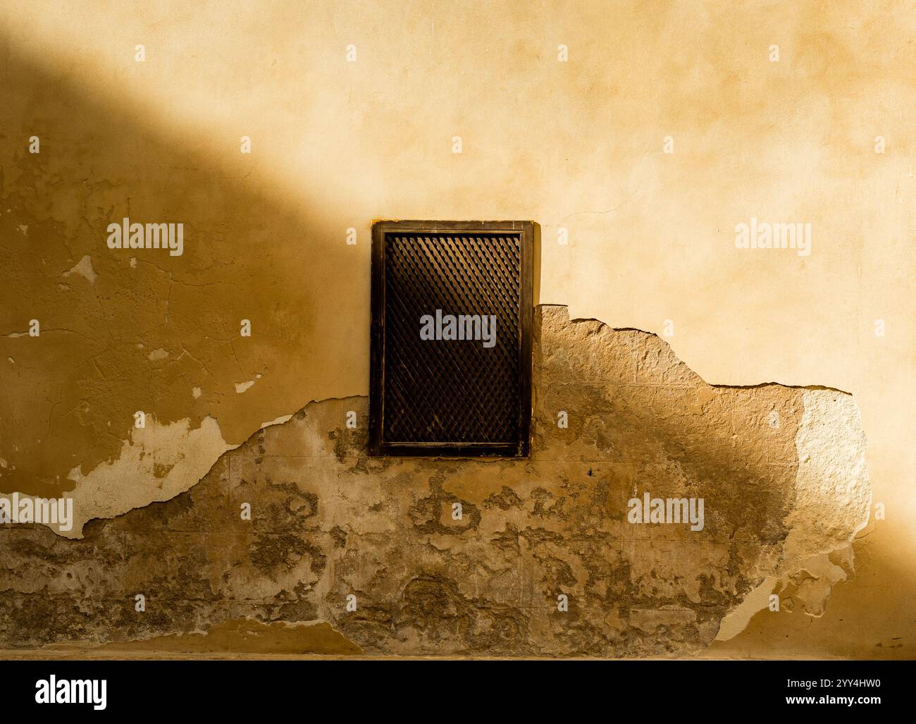 Textured old wall with peeling plaster and a small diamond-patterned window illuminated by soft sunlight and shadow Stock Photo