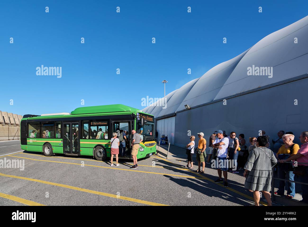 Civitavecchia. Italia - 19 de diciembre de 2024: Pasajeros esperando en fila junto a un autob s verde en una parada en un d a soleado, siguiendo la l Stock Photo