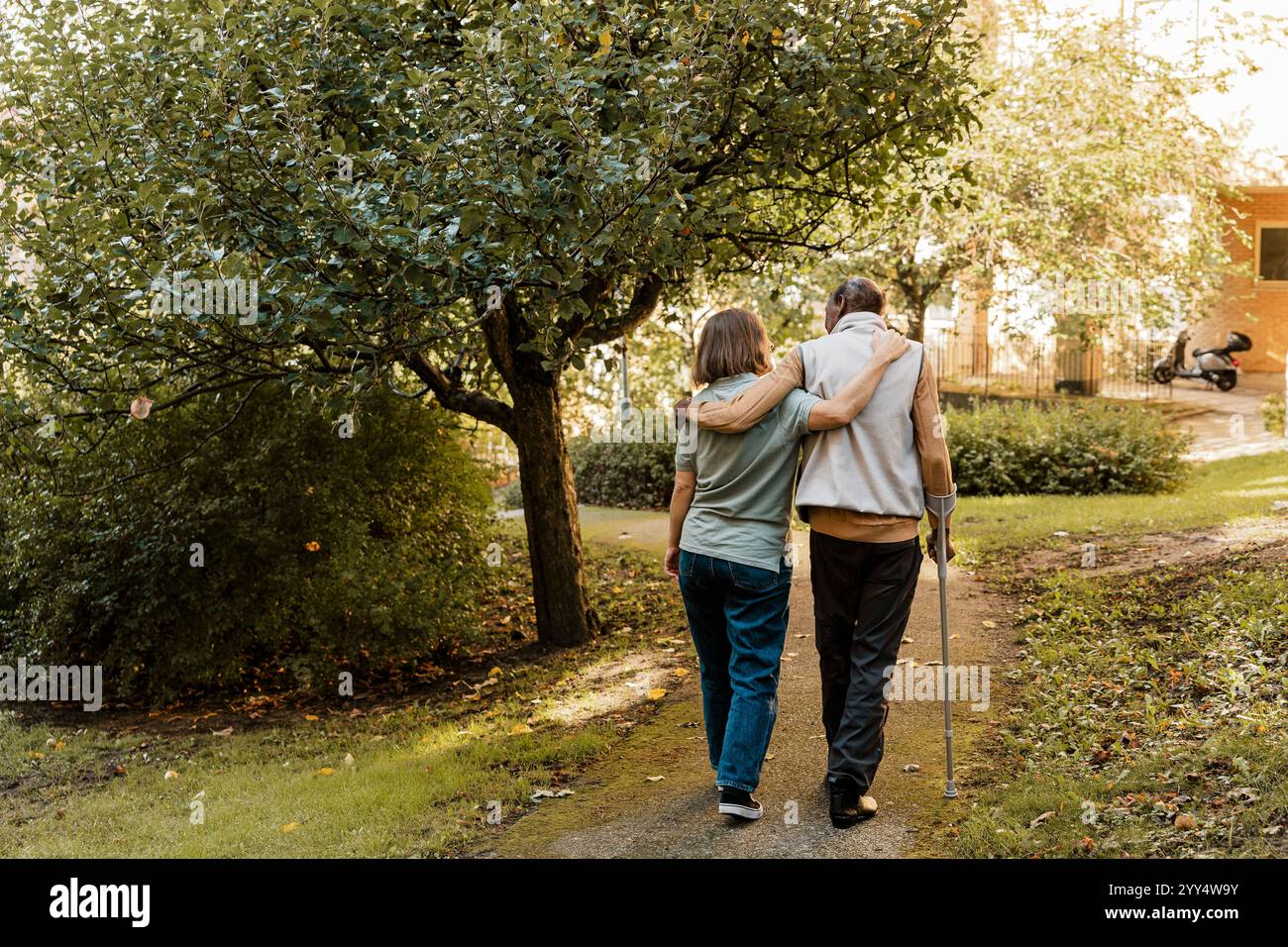 Rear view of female nurse walking with arm around elderly male at park ...