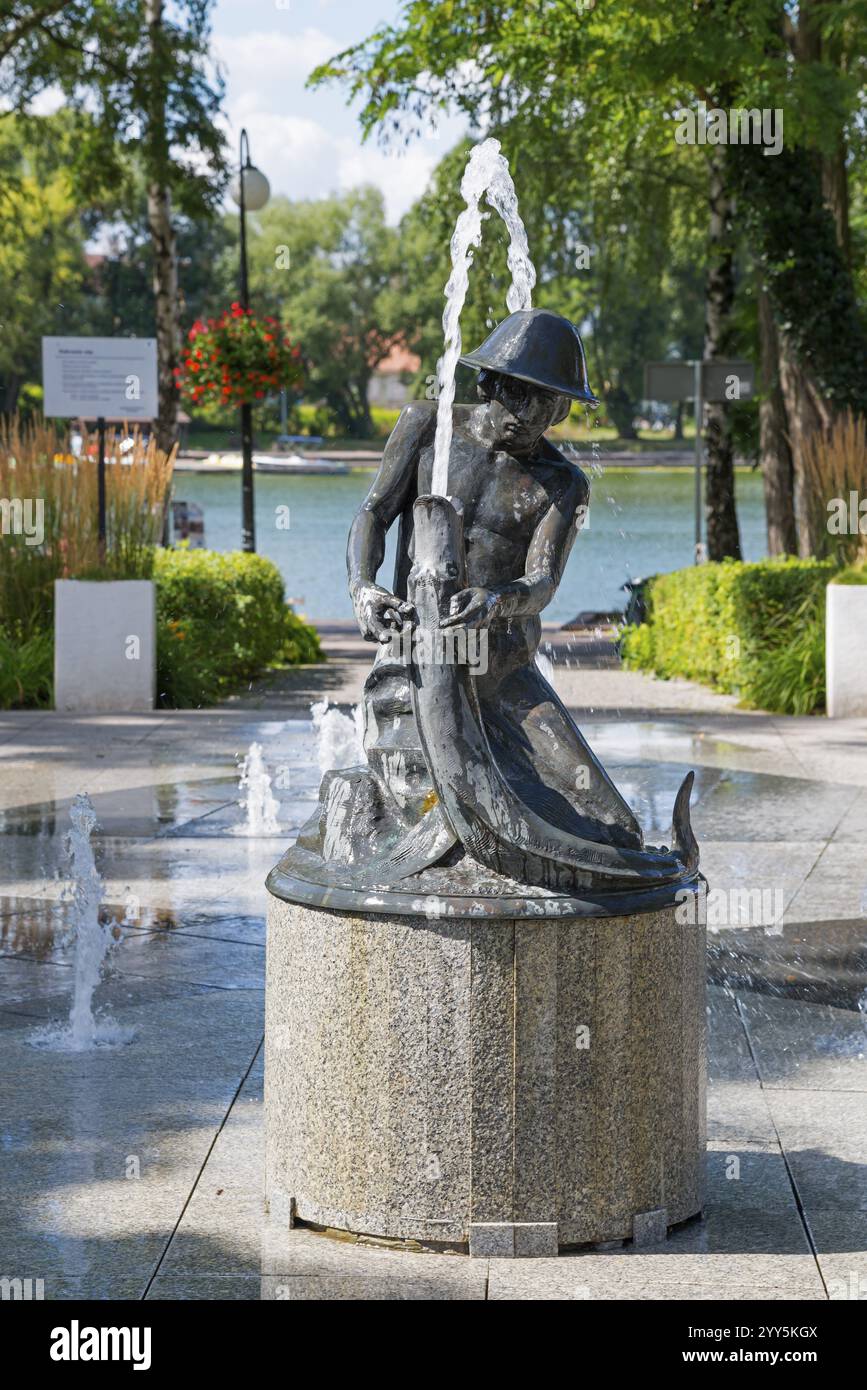 Fountain with statue in a sunny park with trees and water in the background, Ostroda, Osterode in East Prussia, Warminsko-Mazurskie, Warmia-Mazurskie Stock Photo