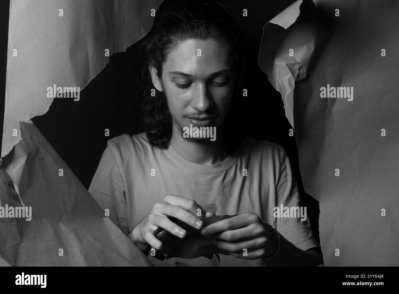 1.126 / 5.000 A man peering through a hole in a sheet of brown paper. Tearing the paper with his hands. Black and white studio portrait. Stock Photo