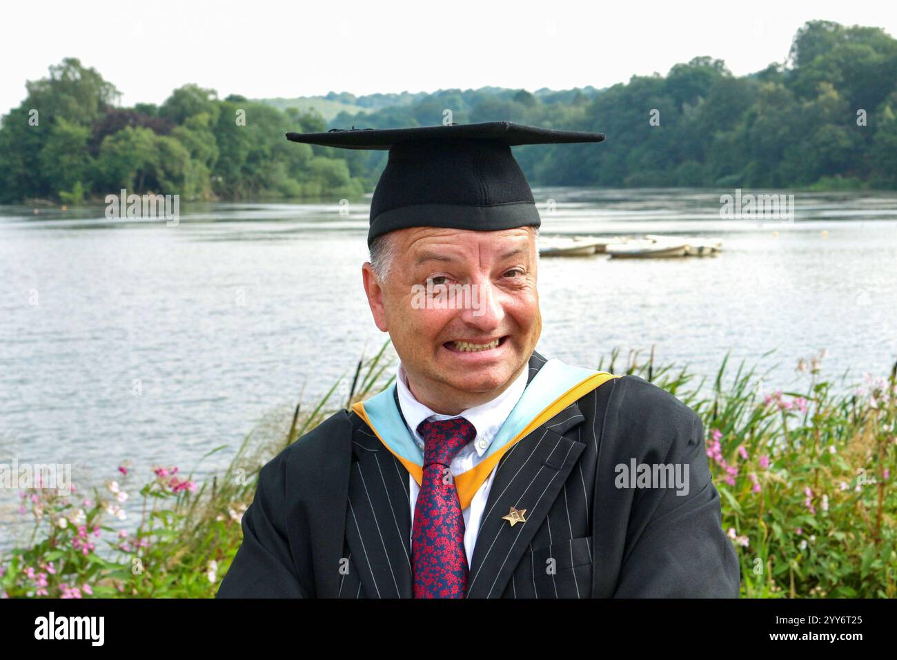 middle aged man in graduation formal dress,  mortar board and gown in front of lake smiling and pulling funny goofy face Stock Photo