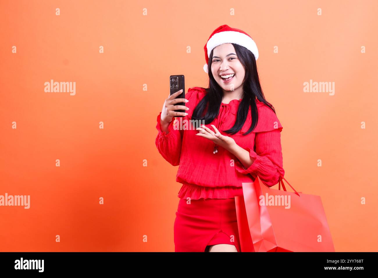 beautiful women charm wearing christmas dress, santa hat, cheerful looking at camera, hand sign present and holding device, carrying red bag of christ Stock Photo