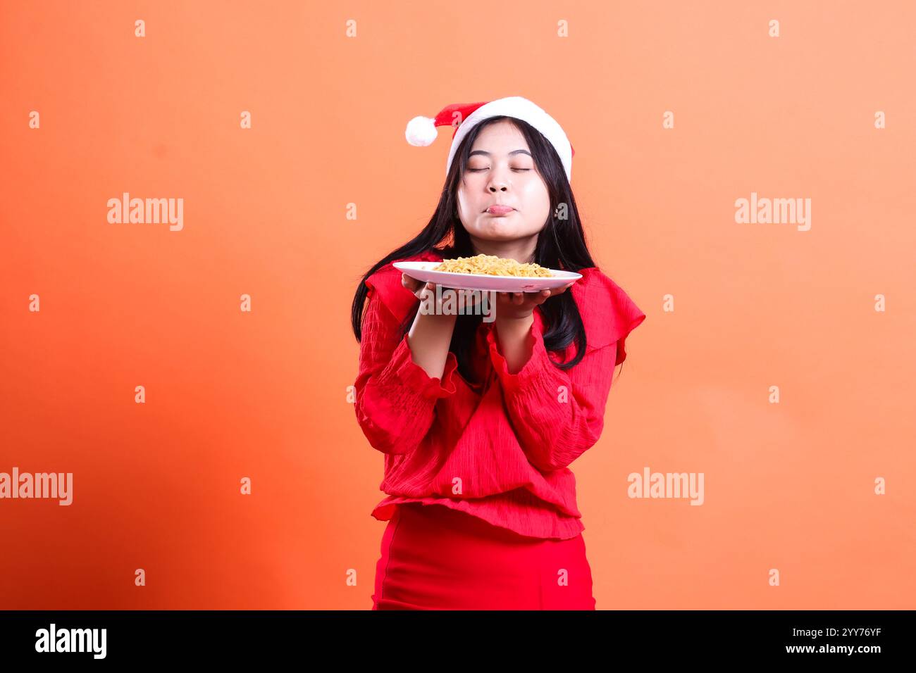 expression of woman wearing christmas dress, Santa hat, eyes closed inhaling the delicious smell of noodles, both hands holding white plate filled wit Stock Photo