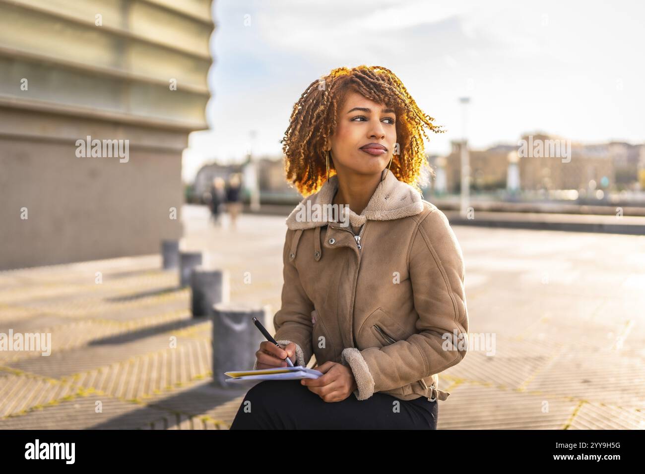 Beauty latin woman with curly hair writing notes sitting in the city during a cold and sunny day Stock Photo