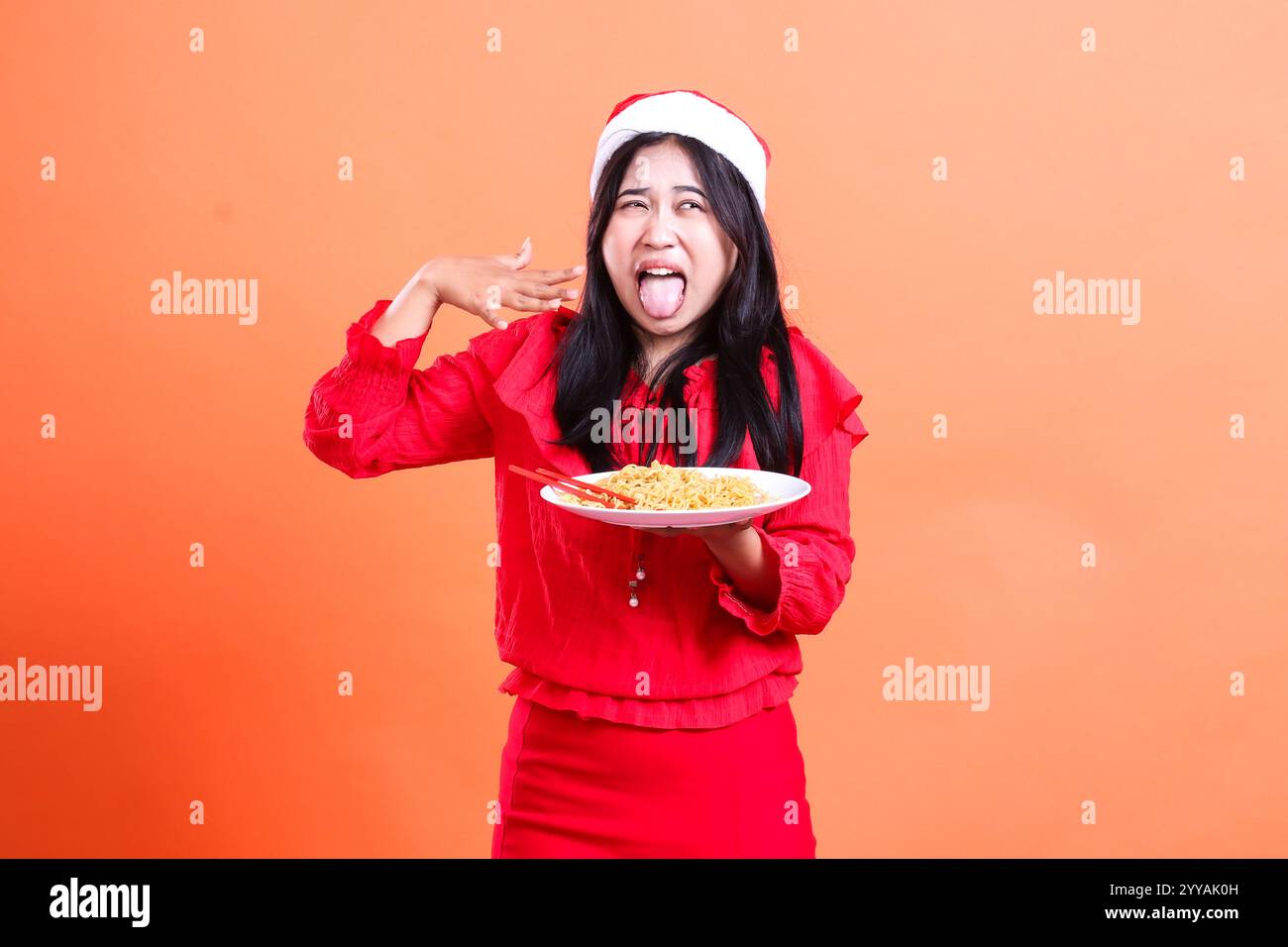 portrait of female wearing christmas blouse, Santa hat, tongue out very spicy eating noodles left eye, hand fanning to mouth and holding white plate f Stock Photo