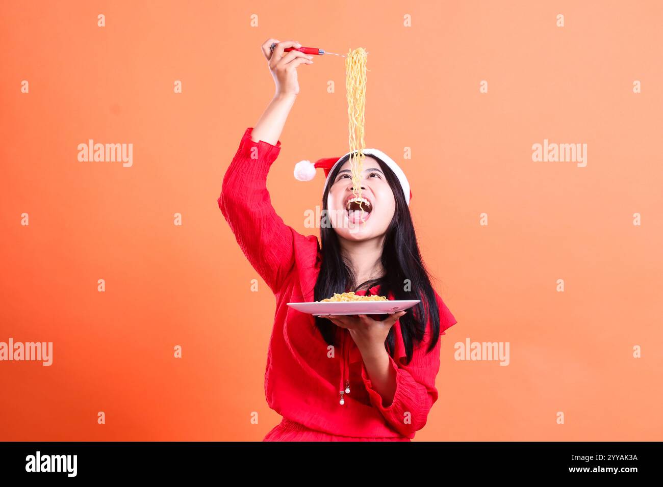 portrait of girl wearing christmas blouse, santa hat, eating noodles see noodles above, hand holding fork filled with noodles above head and white pla Stock Photo