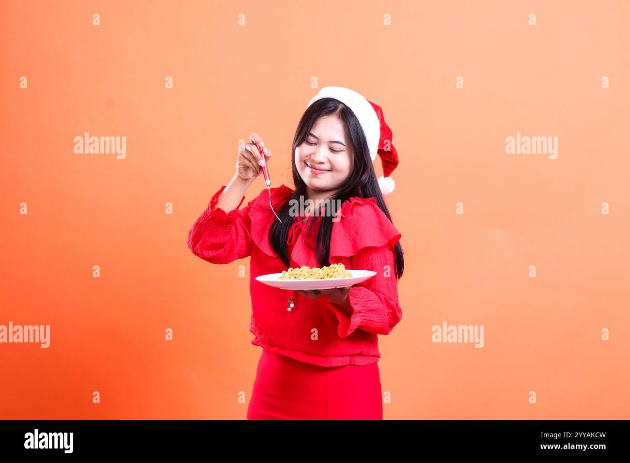 young asian women smiling looking at noodles, hands holding fork and white plate filled with noodles, wearing red christmas blouse, Santa hat, isolate Stock Photo