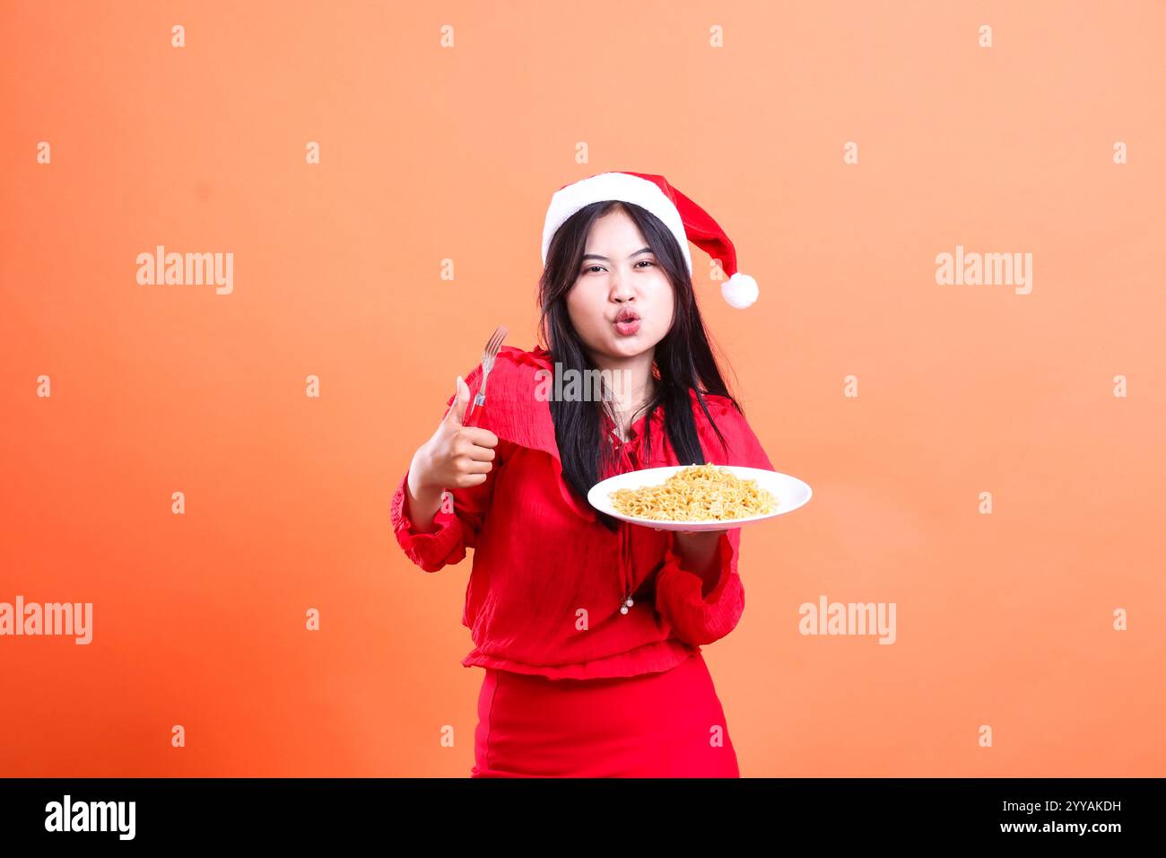 charm of beautiful girl wearing dress, santa hat, kiss looking at camera, hand holding fork while and white plate filled with noodles while thumbs up Stock Photo