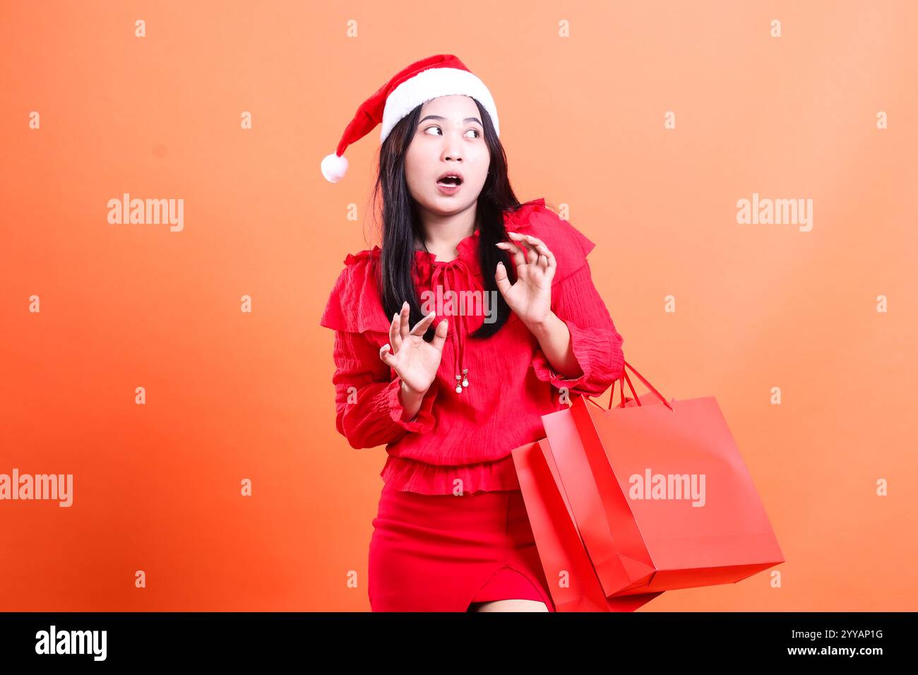 woman wearing red dress and santa hat, standing surprised to the left hand carrying red bag of christmas gifts and surprised sign, wearing red blouse Stock Photo