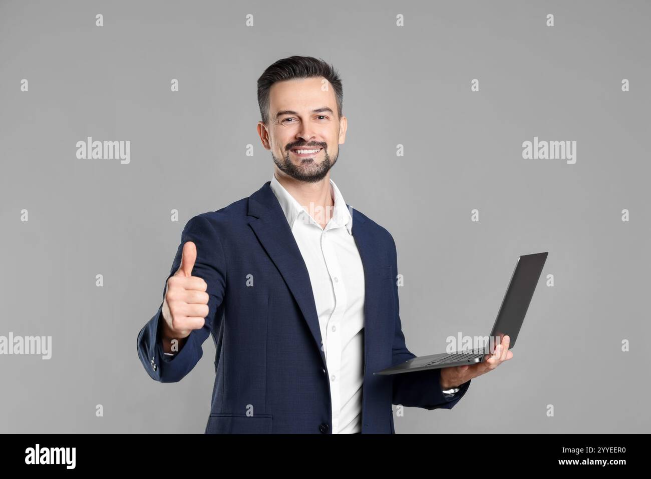 Portrait of smiling banker with laptop showing thumbs up on grey ...