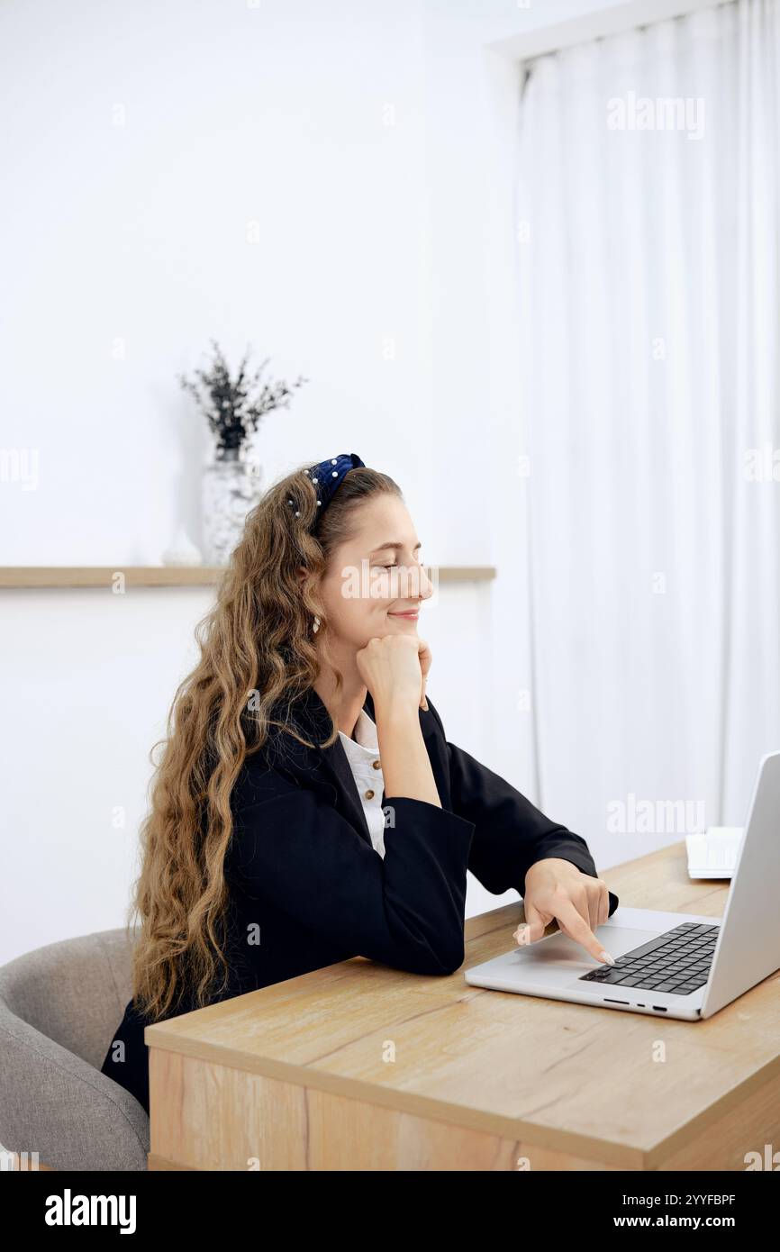 A woman with wavy hair sits at a wooden desk, focused on her laptop ...