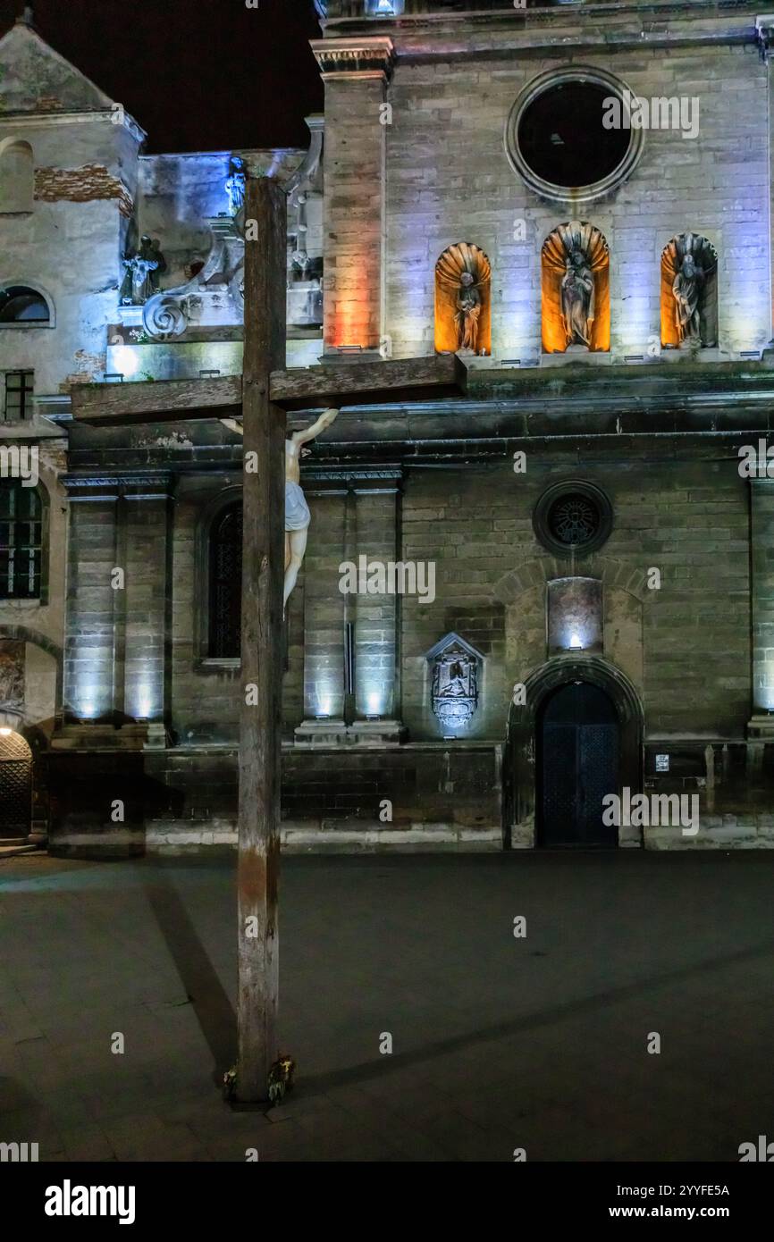 A wooden cross is standing in front of a church. The cross is lit up and the church is lit up as well. The scene is dark and mysterious Stock Photo