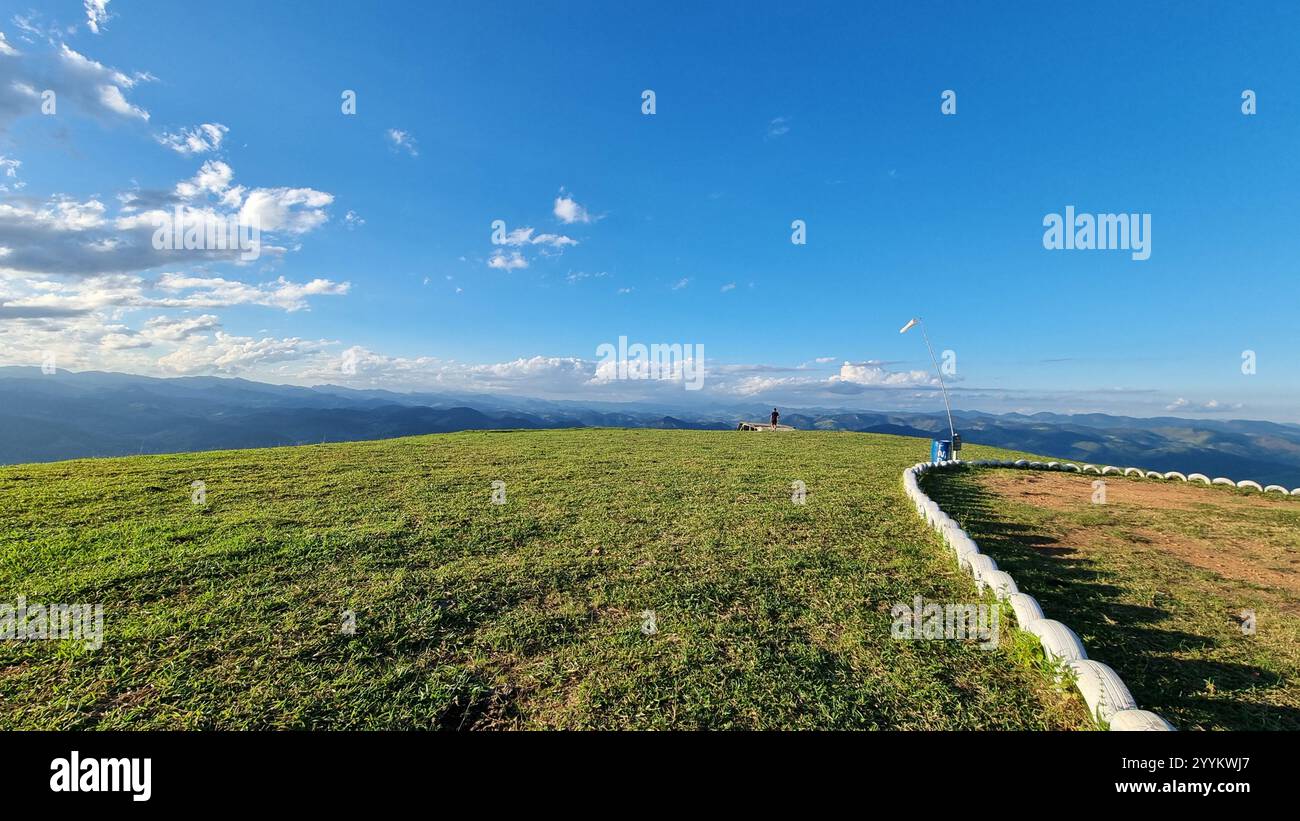 This breathtaking photo captures a serene hilltop with a lush green grass field stretching out under a vibrant blue sky dotted with soft, scattered cl - Smartphone Captured Stock Image