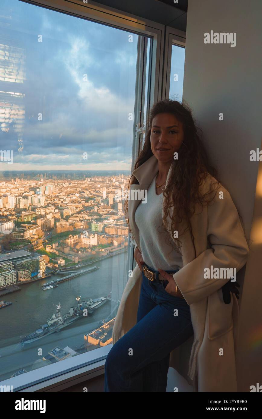 A woman stands by a large window overlooking London's cityscape, featuring the River Thames, Tower of London, and Tower Bridge at sunrise or sunset. Stock Photo