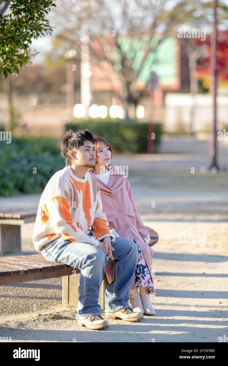 A couple in their 20s are sitting on a bench and talking in a large park in Handa City, Aichi Prefecture in autumn. Stock Photo