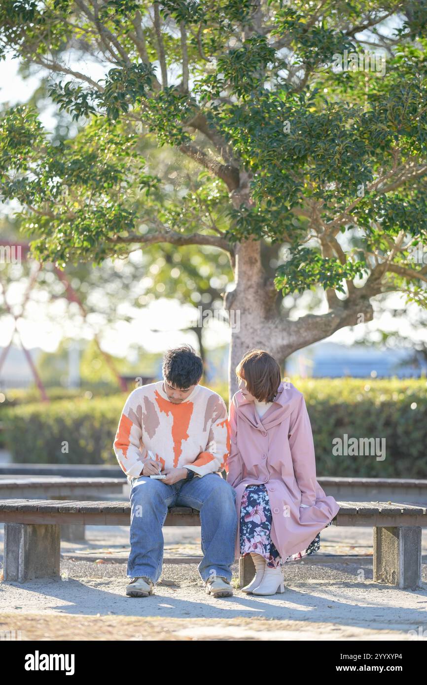 A couple in their 20s are sitting on a bench and talking in a large park in Handa City, Aichi Prefecture in autumn. Stock Photo