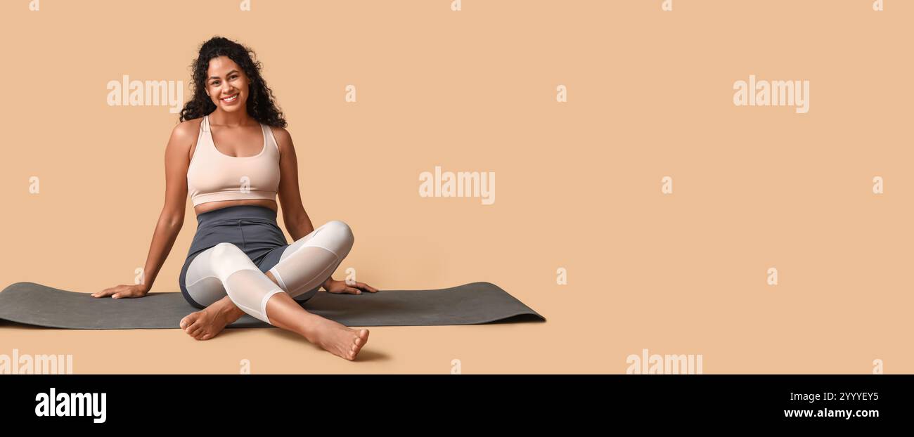 Sporty young African-American woman sitting on yoga mat against beige ...