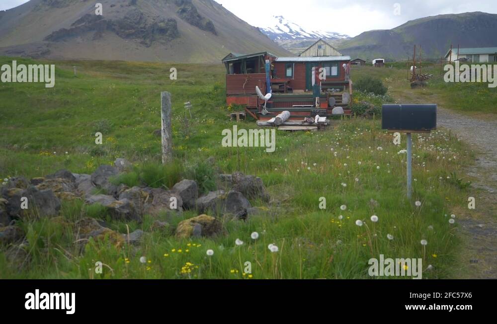 Old run down red and rusty cabin in a green meadow at the base of a snowy Stock Video Footage