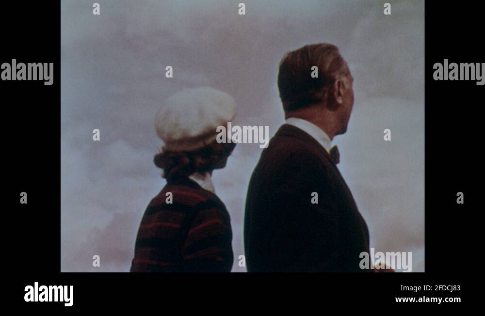 1960s: Man and woman stand looking out over the top of city. Mountain ...