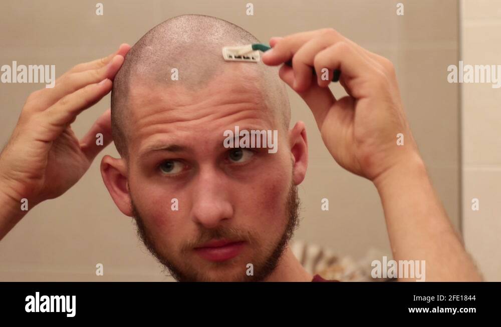 Young bald man shaving his own head looking in the mirror with