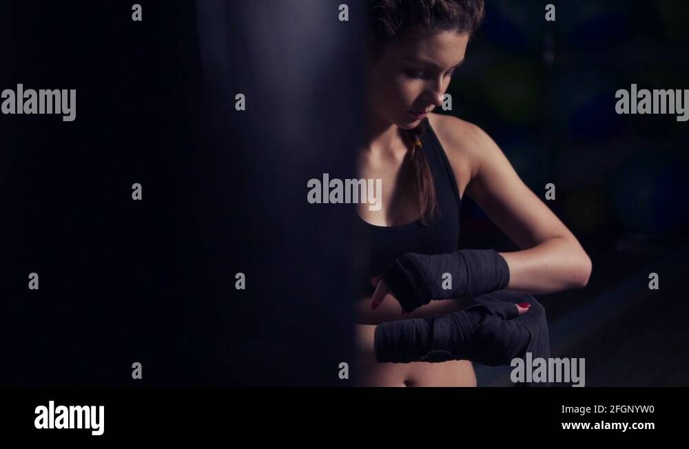 Young beautiful woman wrapping hands with black boxing wraps in dark
