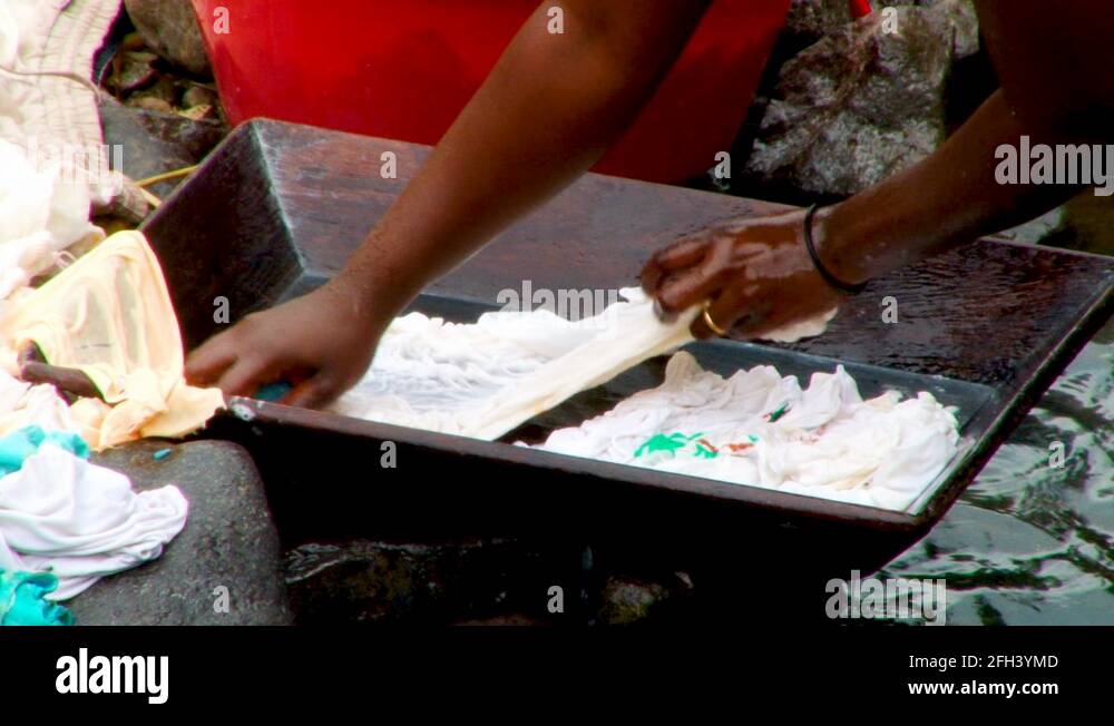 African woman uses a small wooden bin and a rock to wash clothing in a ...