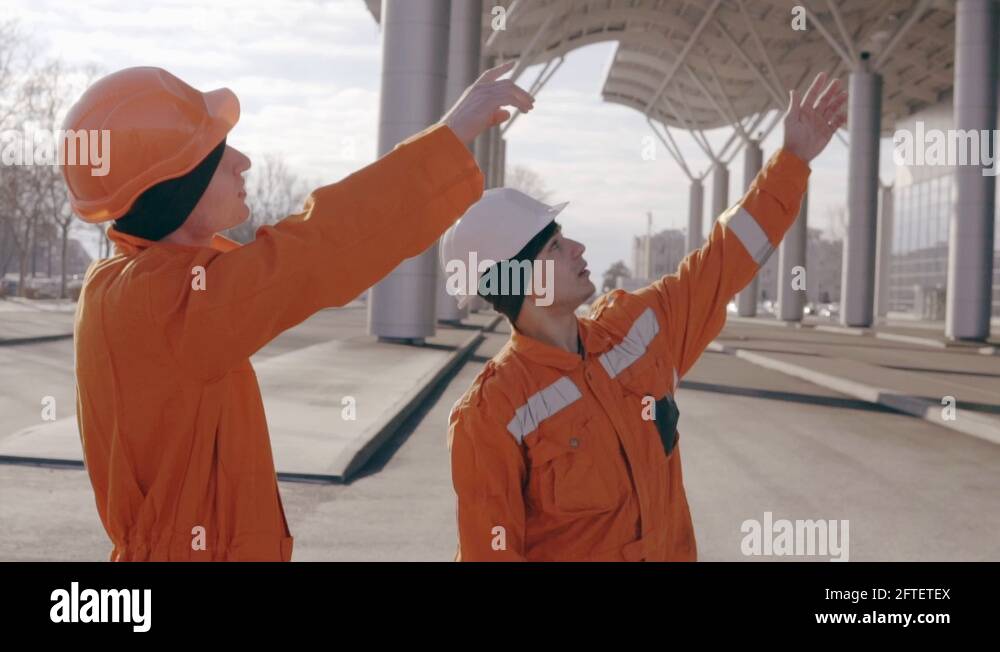 Two workers in orange uniform and helmet pointing at the building and ...