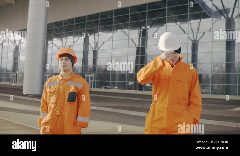 Two construction workers in orange uniform and helmets walking tohether ...