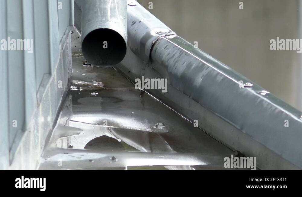 Rainwater dripping out of a steel rain pipe creating rain puddles on a