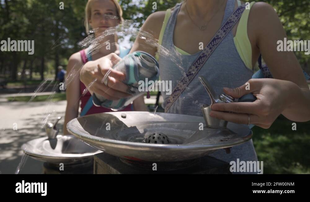 Young Woman Tries To Fill Up Her Water Bottle At A Fountain, Her Friend Laughs Stock Video