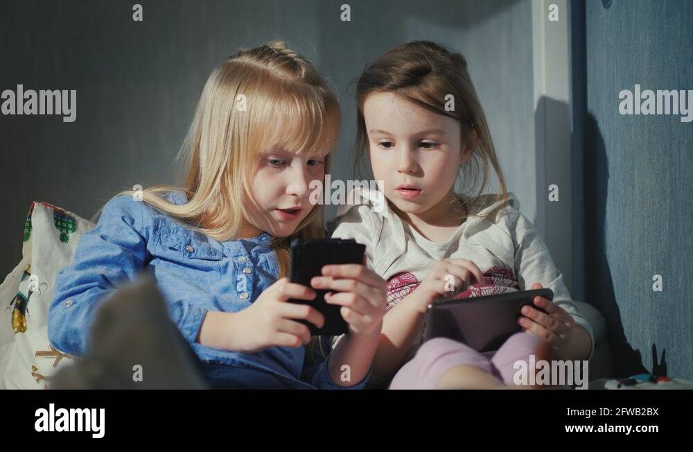 Two girls play on smartphones. Sit side by side on the bed in your