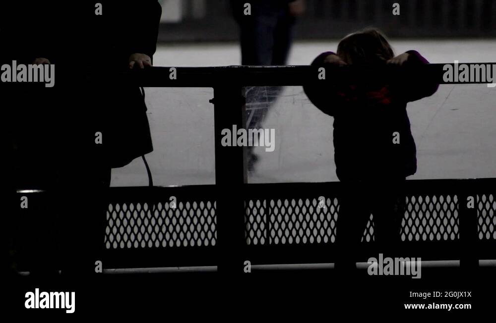 Little girl holds the railing of an outdoor ice skating rink, behind ...