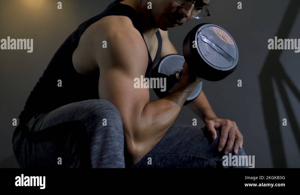 4k Asian man lifting weights dumbbell concentration curl in gym biceps
