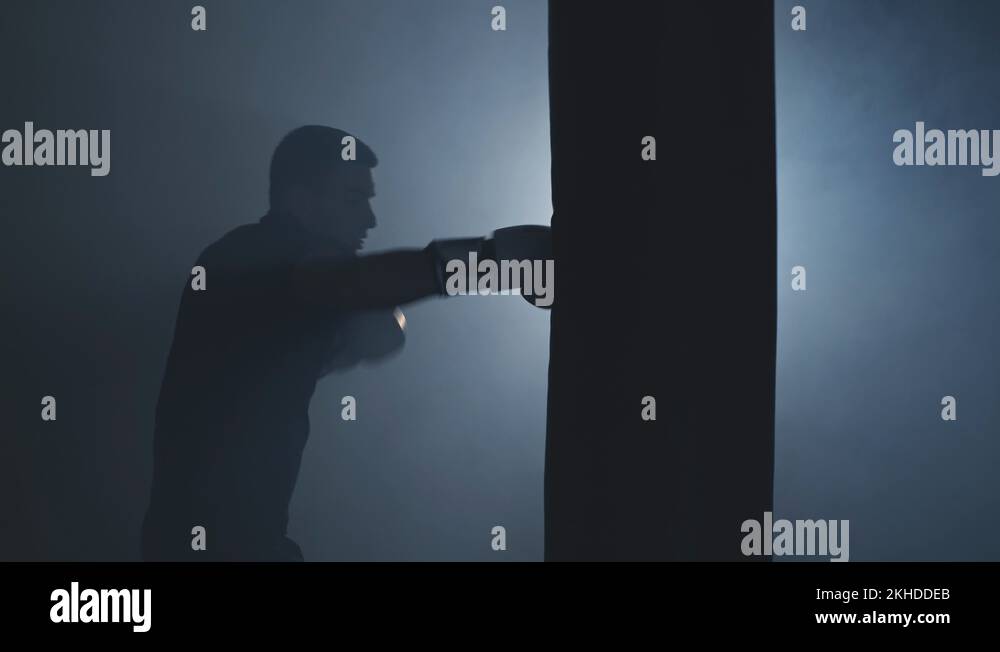 Alone boxer hits punching bag in dark gym. Young man training indoors ...