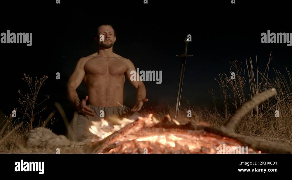 Shirtless male warrior sitting on knees near burning fire with sword