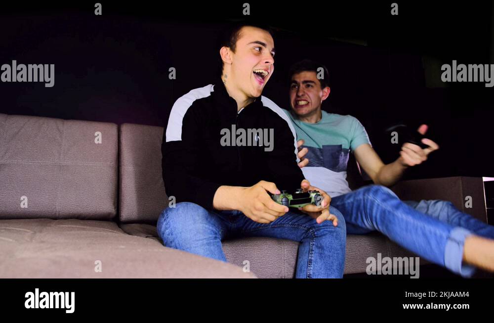 Two young guys laugh. Boys sitting on the couch in front of the TV and ...
