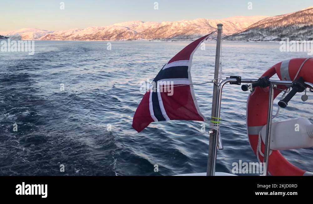 Norwegian flag waving in the wind on the stern of a boat with mountains ...