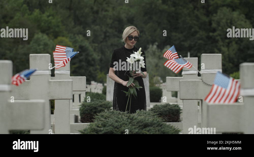 Woman putting white lily flower on grave at cemetery. Widow in dark