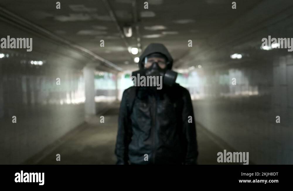 man in gas mask, black jacket with hood standing in long tunnel