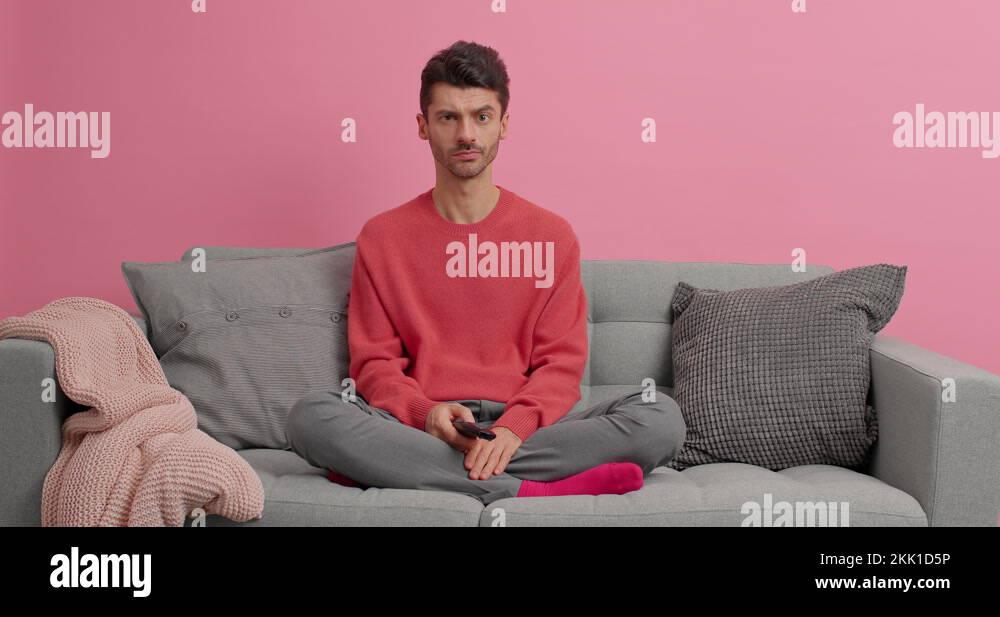 Emotional European man sits crossed legs on sofa with cushions watches  sport game supports favorite Stock Photo by wayhomestudioo